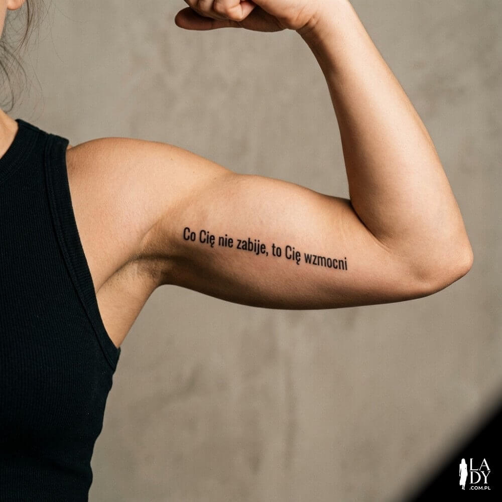 Napis „Co Cię nie zabije, to Cię wzmocni” jako tatuaż na ramieniu kobiety w miękkim świetle studyjnym