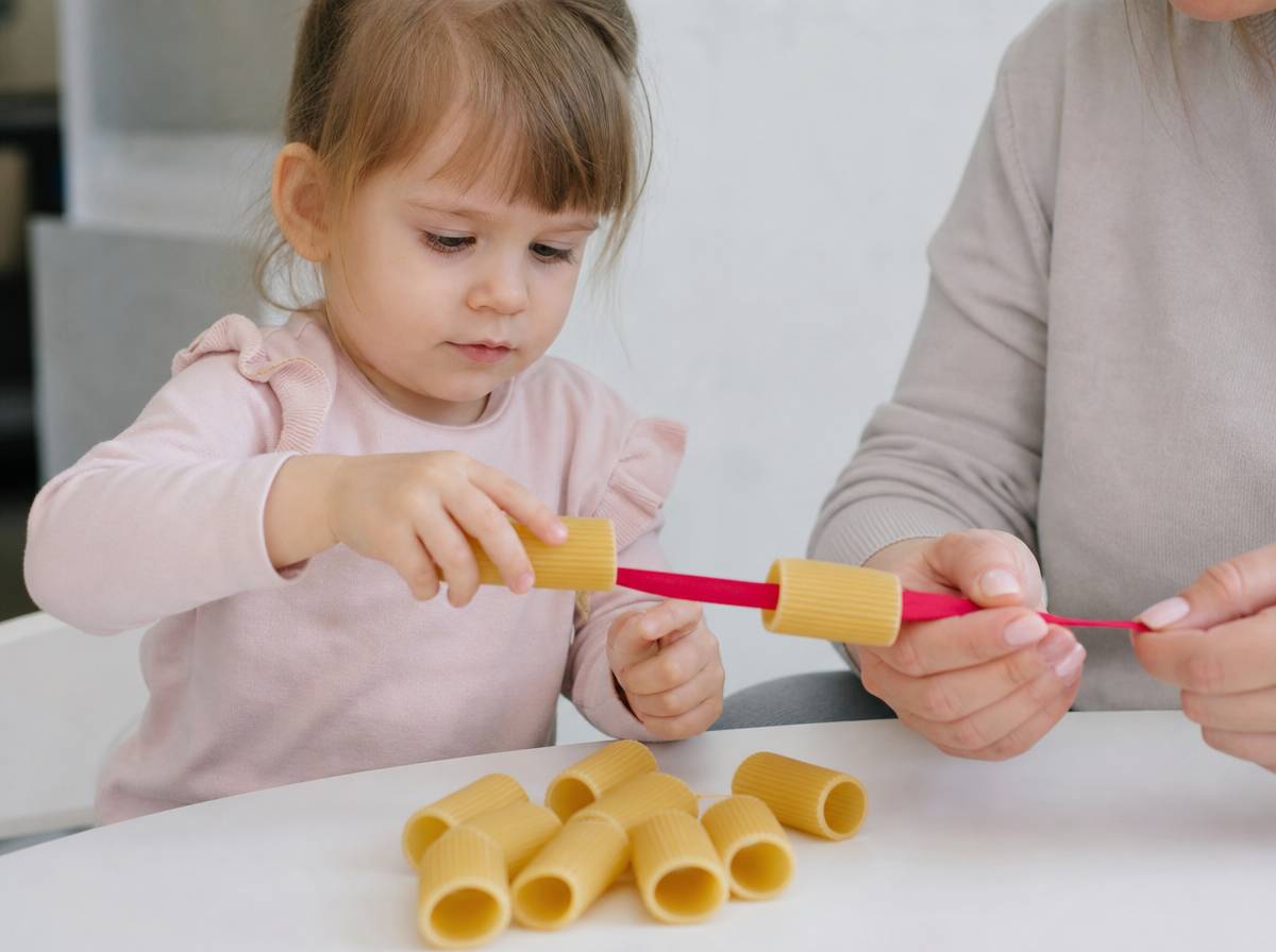 Dziecko nawleka duże rurki makaronu na sztywną tasiemkę trzymaną przez dorosłego